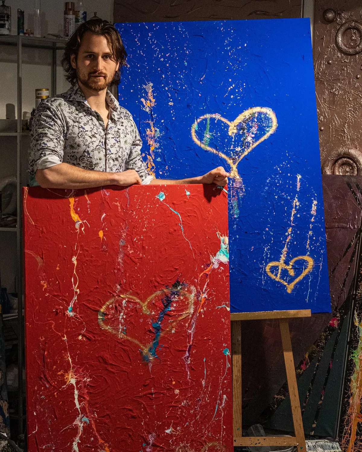 Bis vor kurzem lebte Hojas in Hinwil. Dort hatte er in er Wohnung sein eigenes Studio. AUf dem Foto sieht man einen Mann, der neben einer Leindwand steht. Dort hat es ein blaues Bild. Vor ihm sieht man ein rotes Bild.